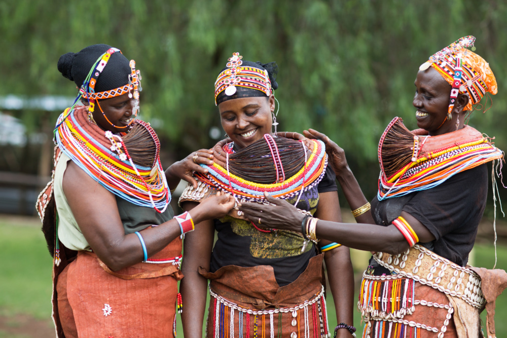 three Indigenous women from Kenya smile while adjusting and sewing beads on to the center woman's traditional clothing