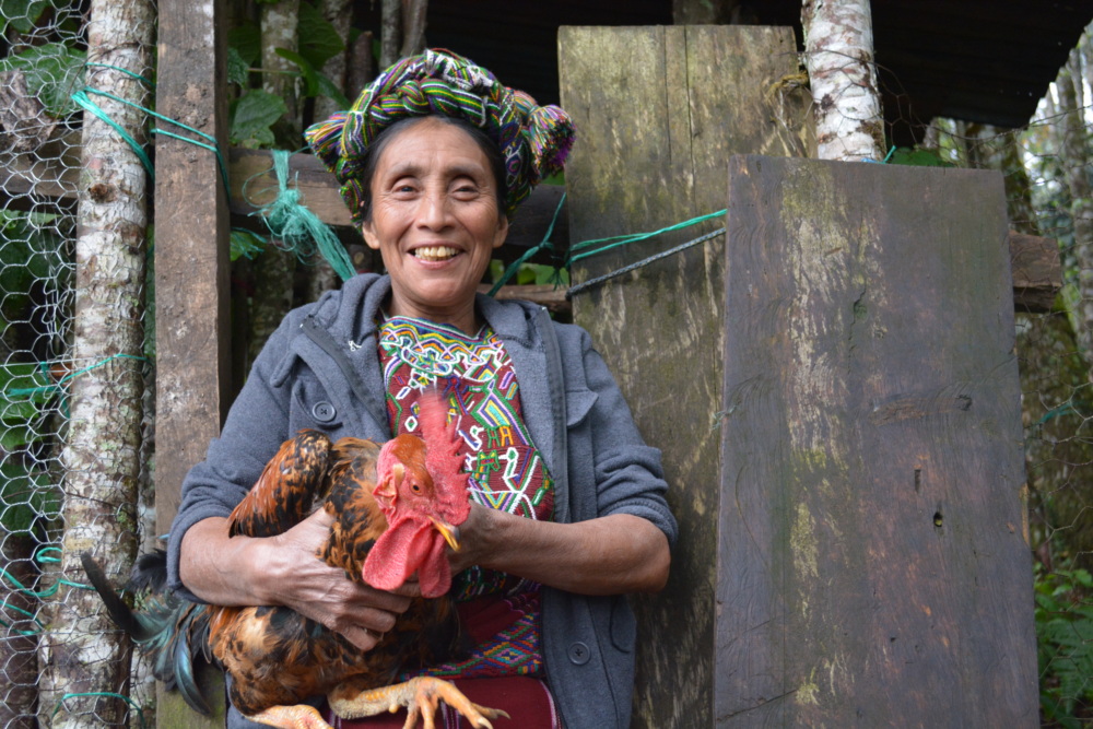 an older Indigenous woman smiles at the camera holding a chicken