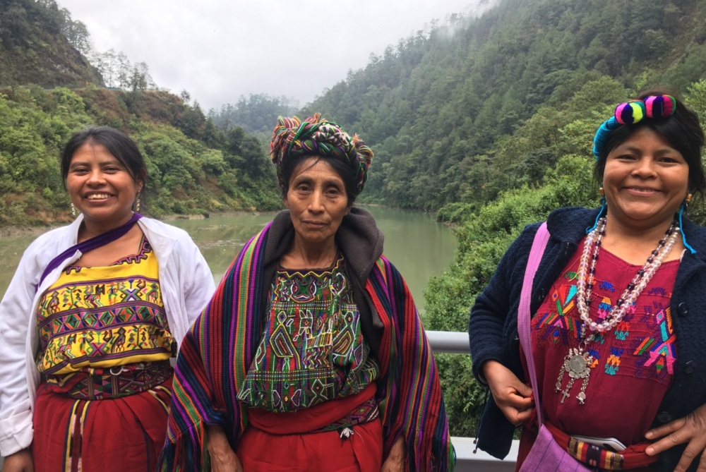 three Indigenous Guatemalan women stand on a bridge over looking a river