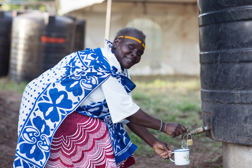 an Indigenous woman in Kenya is bent over pouring water from a water tank