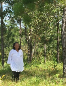 Rose Cunningham Kain wears a long white shirt, with her hair to her shoulders, as she stands amongst the greenery os a natural space.